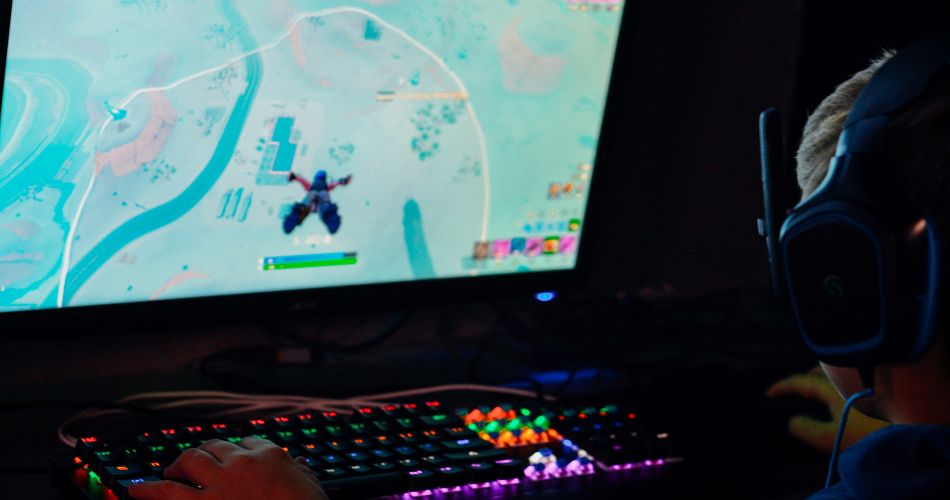 A gamer wearing a headset plays a battle royale game on a PC monitor using a colorful, RGB backlit keyboard in a dark room.