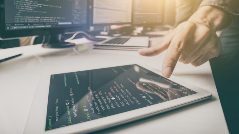 A developer pointing at a tablet screen displaying HTML5 code, with additional monitors and laptops running coding environments in the background. The setup represents the innovation and flexibility of HTML5 in enabling cross-platform gaming.